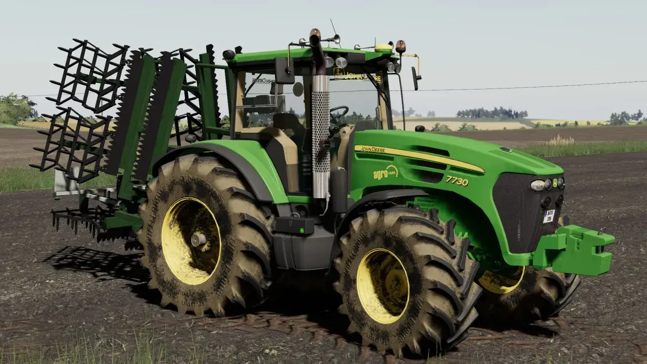 Zielony traktor John Deere 7730 z agregatem uprawowym, gotowy do pracy w polu w grze Ursus FS19.