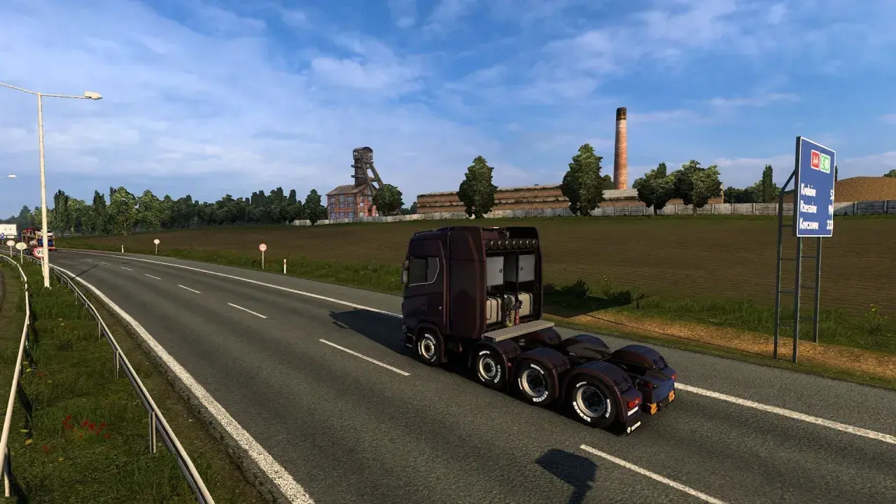 Ciemnobrązowa ciężarówka jedzie na wschód autostradą obok kopalni i pola.