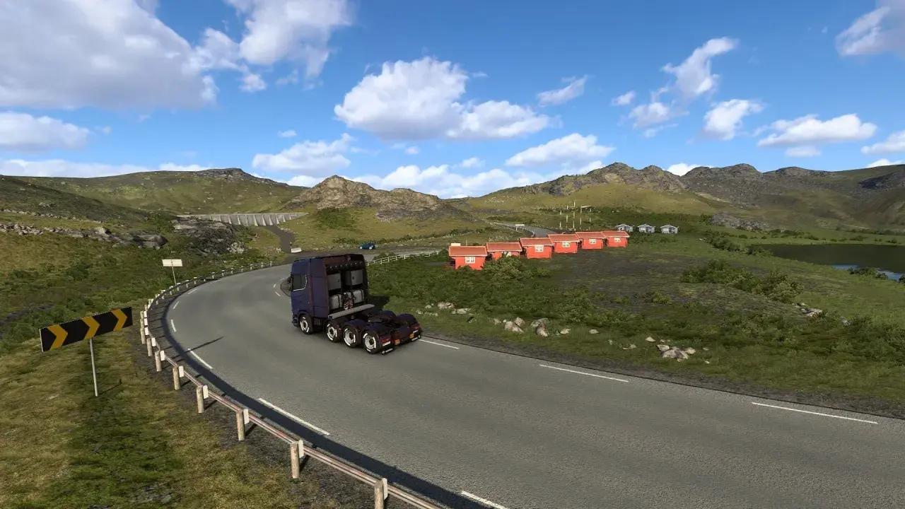 Ciężarówka jedzie krętą drogą w górach. W oddali widać małe czerwone domki i jezioro. To krajobraz z gry ETS2, prawdopodobnie z DLC.
