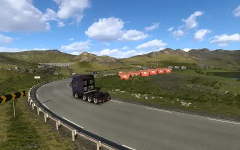 Ciężarówka jedzie krętą drogą w górach. W oddali widać małe czerwone domki i jezioro. To krajobraz z gry ETS2, prawdopodobnie z DLC.