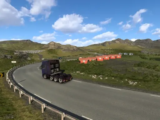 Ciężarówka jedzie krętą drogą w górach. W oddali widać małe czerwone domki i jezioro. To krajobraz z gry ETS2, prawdopodobnie z DLC.