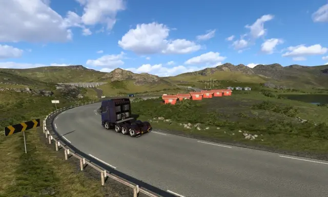 Ciężarówka jedzie krętą drogą w górach. W oddali widać małe czerwone domki i jezioro. To krajobraz z gry ETS2, prawdopodobnie z DLC.